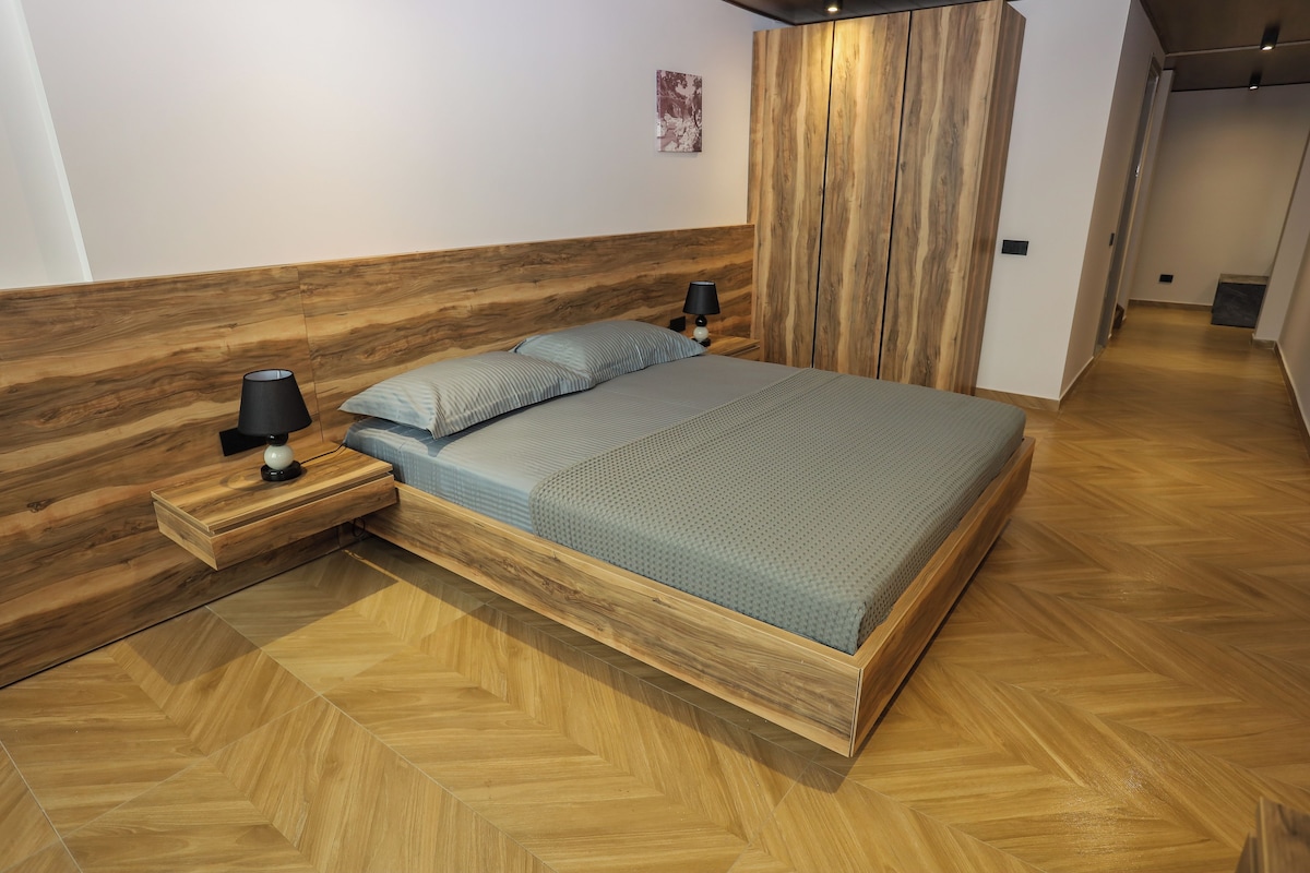 Un dormitorio moderno con cabecera de madera, una cama con colchón gris, dos mesitas de noche con lámparas y un suelo de madera con estampado de espiga.