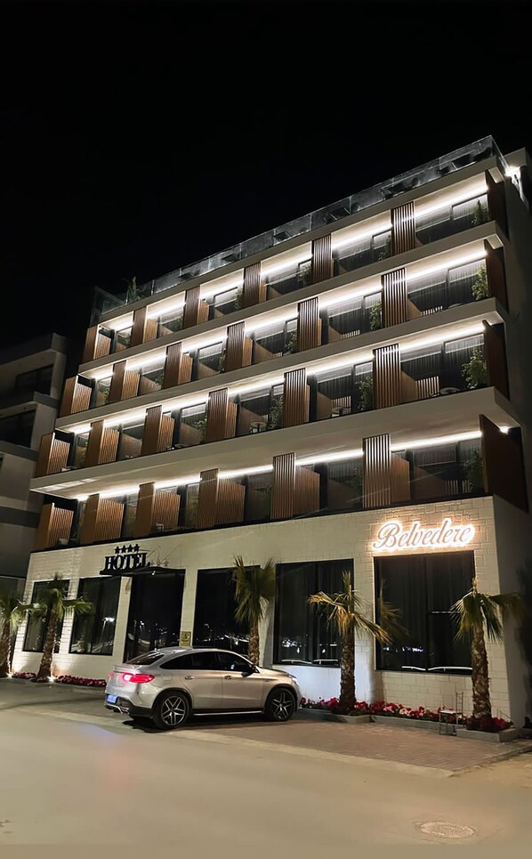 Un hotel de varios pisos con diseño moderno, que cuenta con balcones de listones de madera y un letrero de neón que dice "Hotel Belvedere".