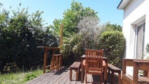 Une terrasse en bois avec une table et des chaises, entourée de verdure et bordée d’un bâtiment blanc.