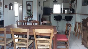 Un coin repas avec une table et des chaises en bois, une cuisine à l’arrière-plan et une plante sur le rebord de la fenêtre.