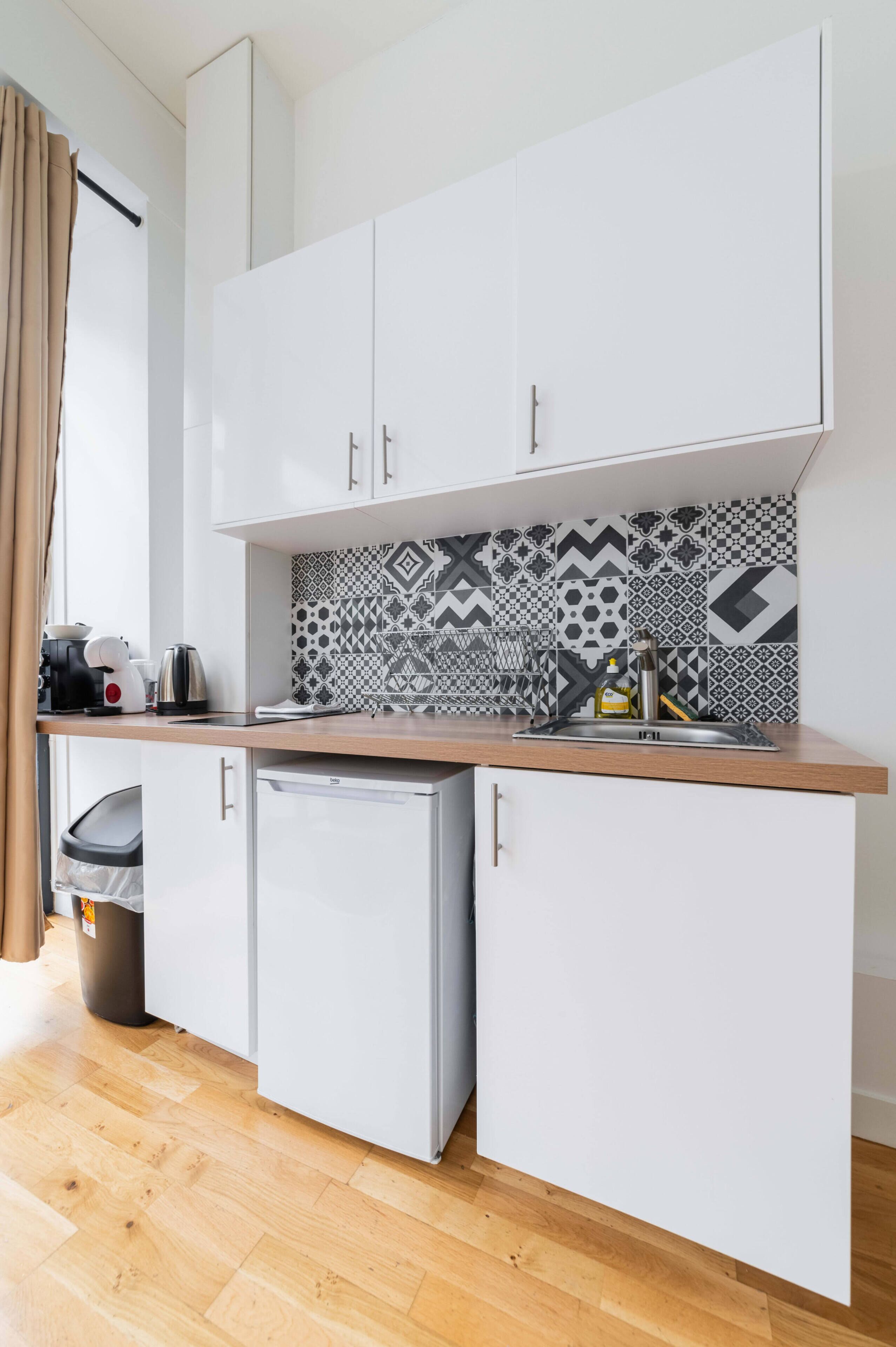 A compact kitchen with white cabinets, a small refrigerator, a patterned backsplash, and a window with curtains.