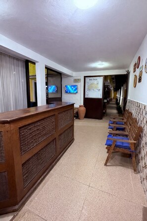 Una zona de recepción con mostrador de madera, dos televisores, un tablón de anuncios y una fila de bancos de madera con cojines azules.