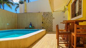 An outdoor swimming pool with a wooden deck, a wooden table and chairs, and a tiled wall with a patterned design. - Pousada Farol do Porto  (Ipojuca)