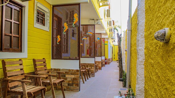 A narrow alley with wooden benches and a yellow building with a window and a door. - Pousada Farol do Porto  (Ipojuca)