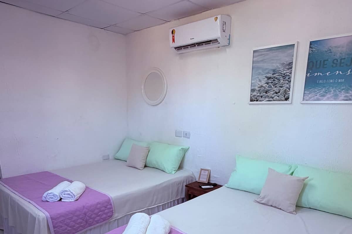 A hotel room with two beds, each with a purple bedspread and white linens, a ceiling-mounted air conditioner, and a wall-mounted round mirror.