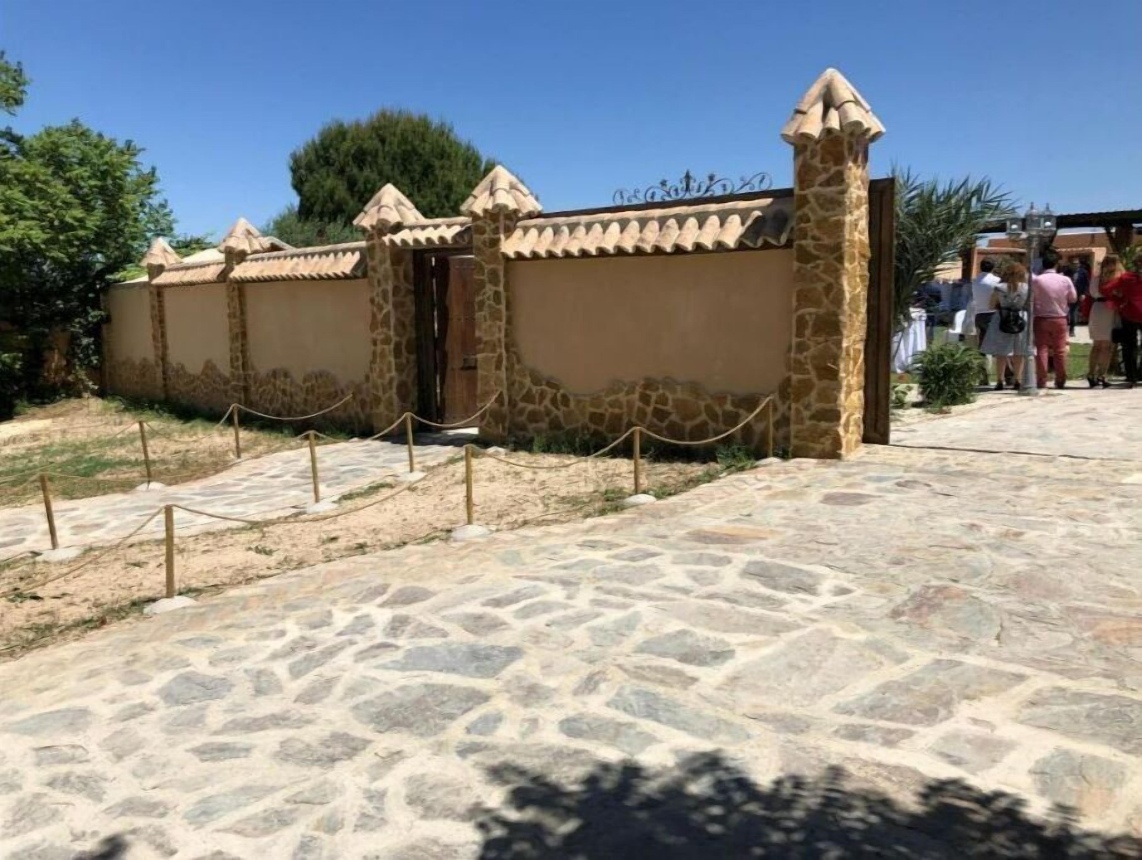 A stone wall with a gate, leading to a paved area with a stone pattern.
