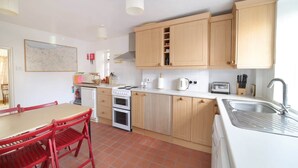 A compact kitchen with wooden cabinets, a white countertop, a red dining table, and a tiled floor. - Beautiful Cottage & Garden, Quantock Hills (Bridgwater)