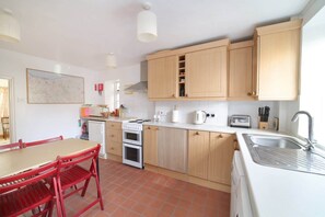 A compact kitchen with wooden cabinets, a white countertop, a red dining table, and a tiled floor. - Beautiful Cottage & Garden, Quantock Hills (Bridgwater)
