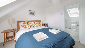 A bedroom with a sloped ceiling, a bed with a blue bedspread, two nightstands with lamps, a picture on the wall, and a view of a balcony with towels.