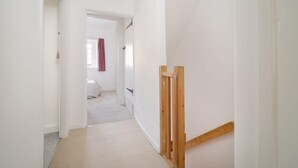 A narrow hallway with white walls and a carpeted floor leading to a bedroom with a bed and a window with a red curtain.