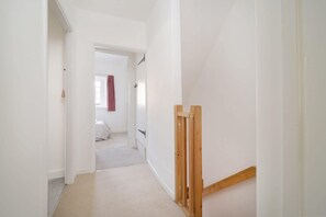 Un couloir étroit aux murs blancs et au sol recouvert d’une moquette mène à une chambre à coucher où se trouvent un lit et une fenêtre ornée d’un rideau rouge.