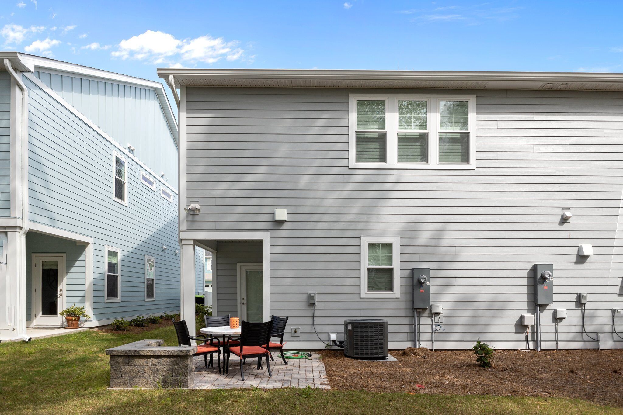 A two-story house with a patio area featuring a table and chairs, outdoor electrical boxes, and a small garden.