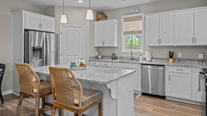 A modern kitchen with white cabinetry, a stainless steel refrigerator, a granite countertop, and two wicker chairs.