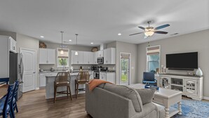 A modern kitchen with white cabinets, a dining area with blue chairs, a gray sofa, and a TV on a white cabinet.