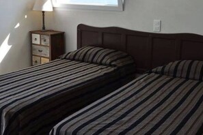 A room with two single beds with striped bedding, a wooden nightstand with a lamp, and a window above the beds.
