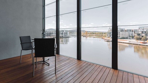 Ein Zimmer mit großen Fenstern, die auf einen Fluss und Gebäude blicken. Es hat einen Holzboden und einen Sessel.