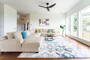 A living room with a sectional sofa, a ceiling fan, a large window, and a floor-to-ceiling view. - Relax & Play in Style: Luxury Home w/ Infinity Pool, Game Room & Alfresco Dining (Jonestown)
