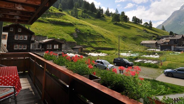 Un balcon en bois avec une table et des chaises rouges, donnant sur une colline verdoyante où l’on aperçoit des bâtiments et des voitures garées.