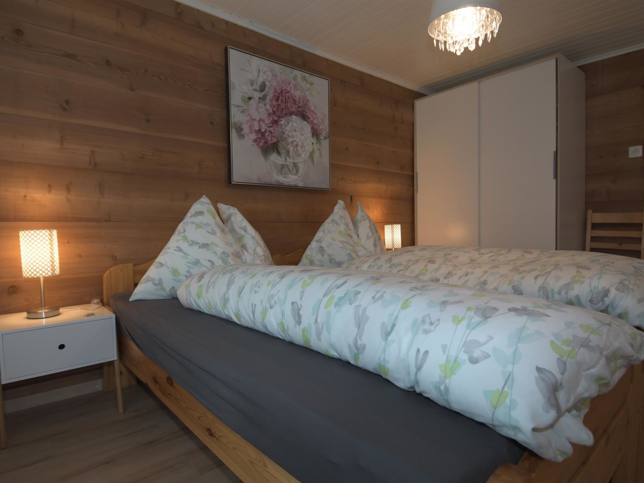 A bedroom with a wooden headboard, a bed with floral bedding, a nightstand with a lamp, and a painting of flowers on the wall.