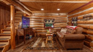 A log cabin interior with a wooden staircase, a television, a sofa, and a rug. - Log Cabin Hot Tub Getaway (Walhonding)