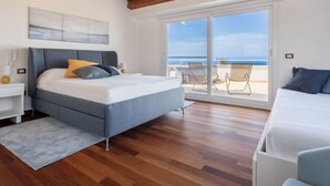 Une chambre à coucher comprenant un lit, une table de chevet, une commode, une grande fenêtre donnant sur la mer et un balcon avec des chaises.