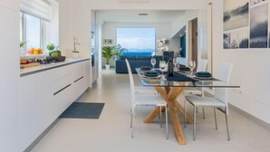 Une cuisine moderne avec un coin repas, comprenant une table en verre et des chaises blanches, une vue sur la mer et une table à manger dressée pour six personnes.