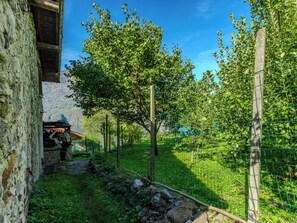 A rustic stone building with a wooden structure attached, surrounded by lush greenery and a clear blue sky.