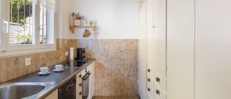 A compact kitchen with a sink, stove, and cabinets. There is a window providing natural light and a small shelf with kitchen items.