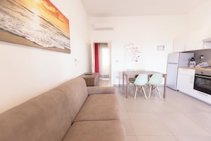 A modern living room with a beige sofa, a dining area with two chairs, and a kitchen with a refrigerator and stove.