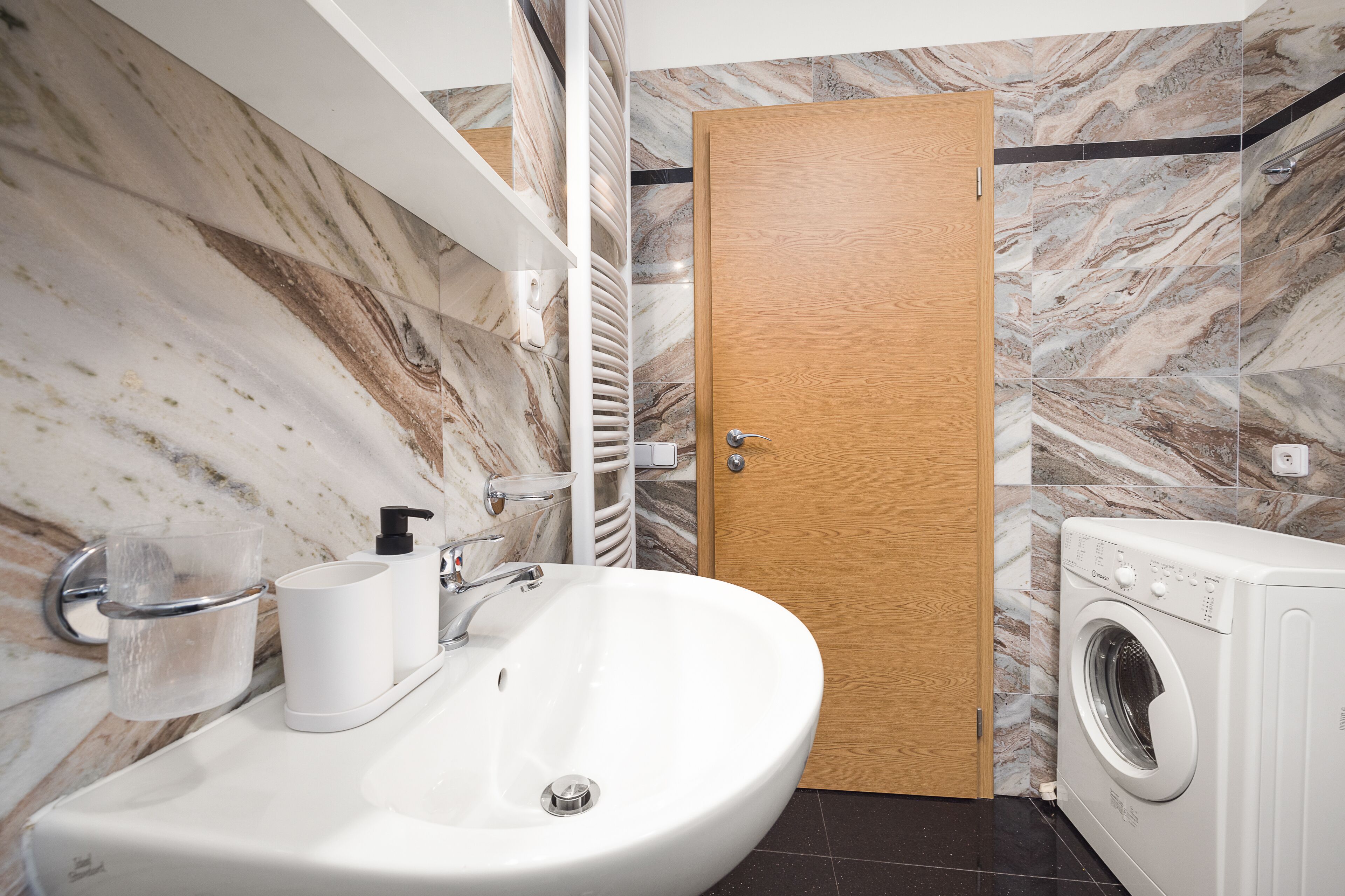 Een badkamer met een marmeren wand, een wastafel, een toiletpapierhouder en een wasmachine.