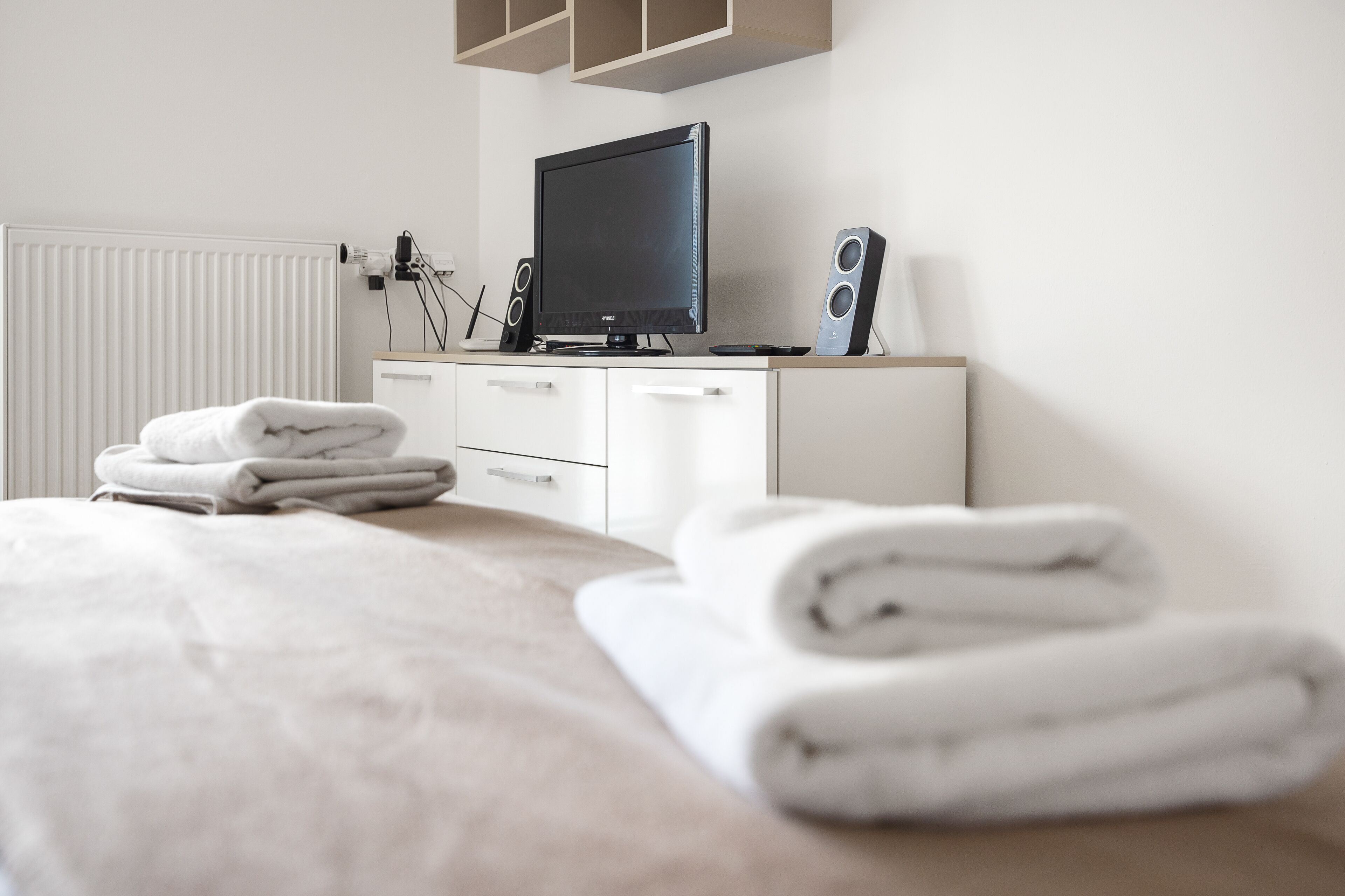 A neatly made bed with white linens and rolled towels, a television on a wooden stand, and a white dresser with drawers.