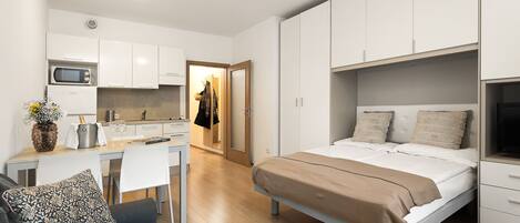 Un dormitorio moderno y funcional con cama, mesitas de noche y una televisión empotrada. Junto a él, una kitchenette con microondas y un pequeño comedor.
