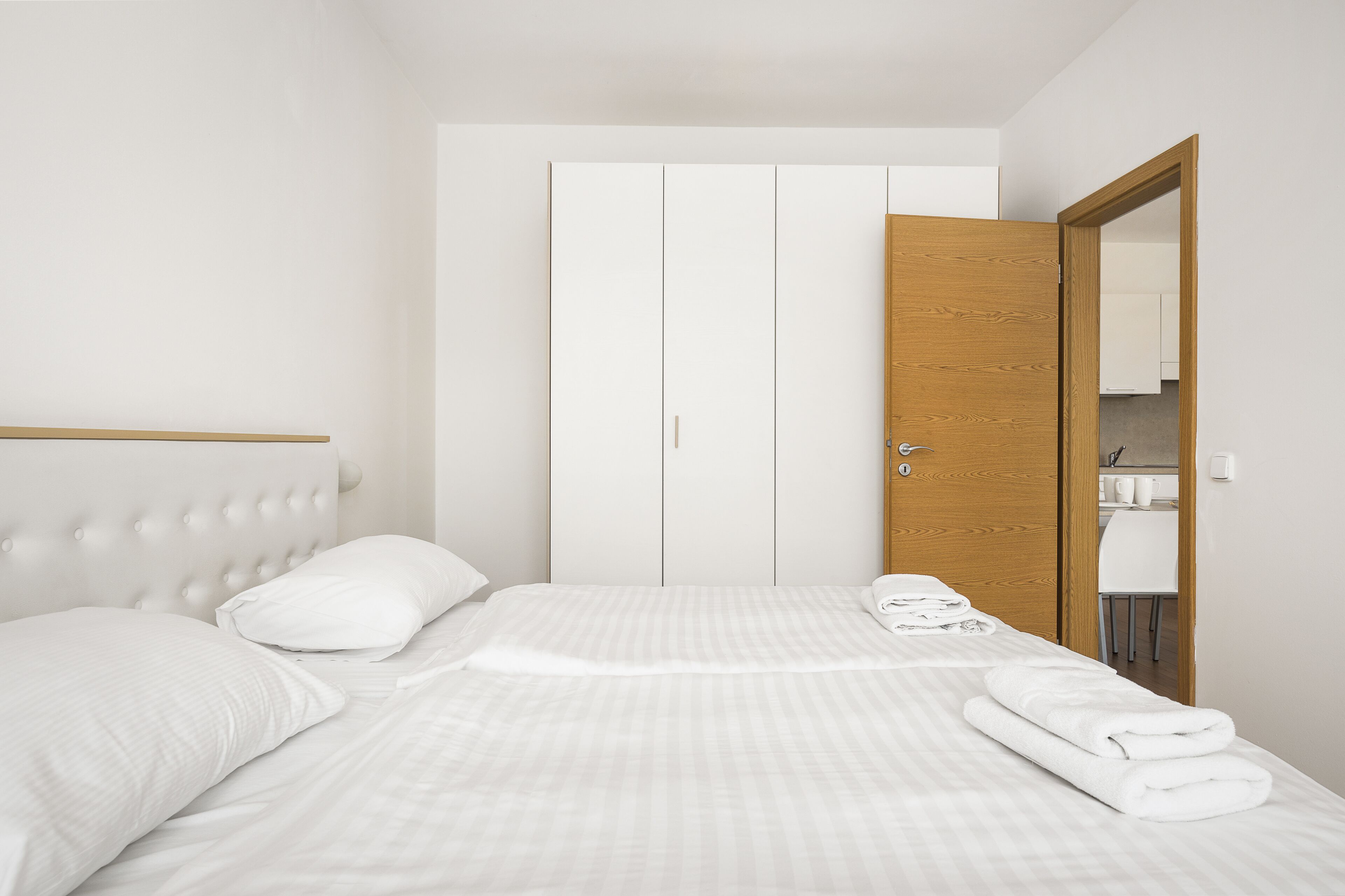 Una cama bien hecha con sábanas blancas, cabecera de madera y un armario blanco con puerta de madera.