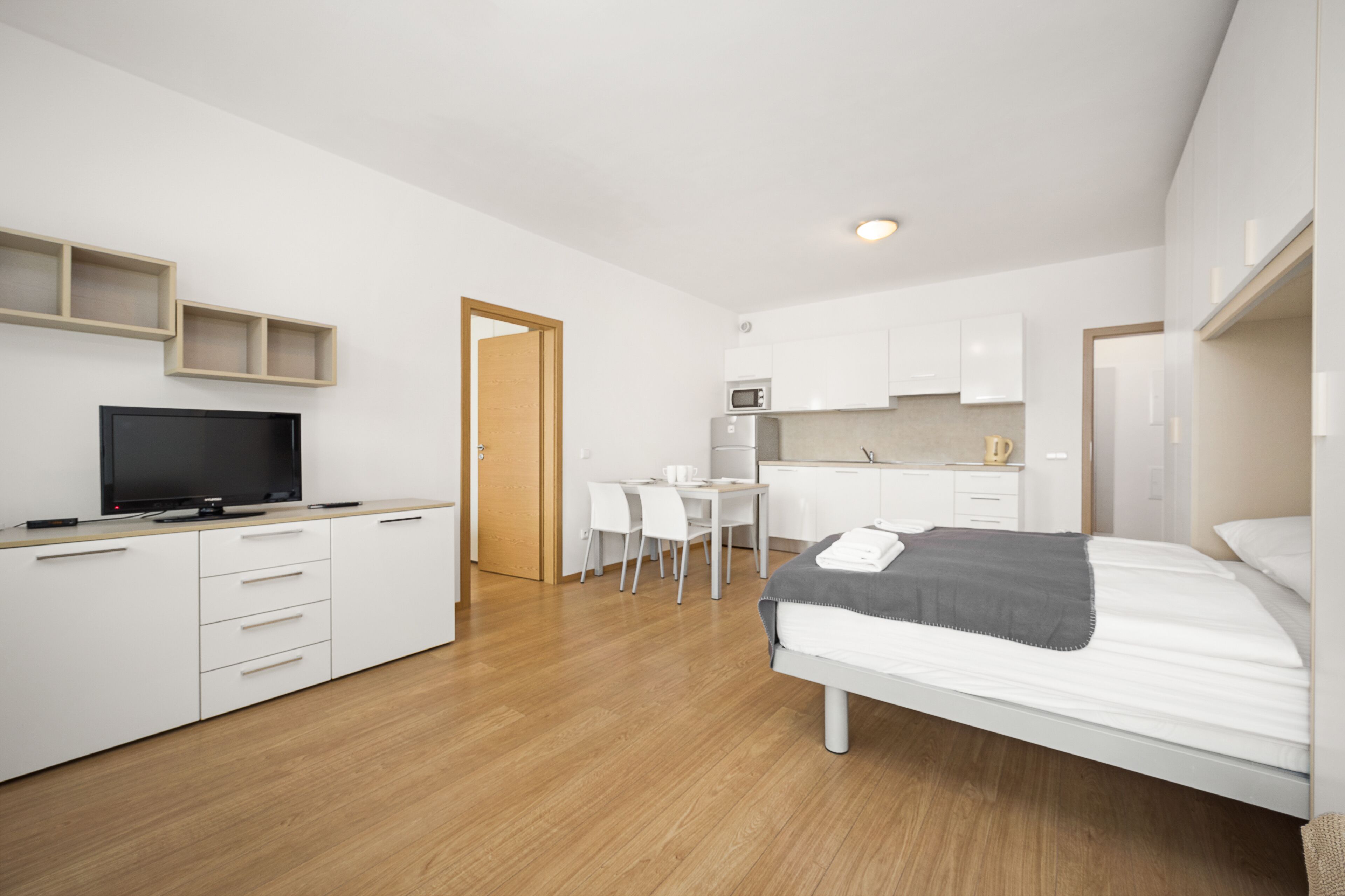 Une chambre moderne et bien éclairée, équipée d’un lit, d’une table de chevet et d’un téléviseur. À côté se trouve une petite cuisine équipée d’un micro-ondes et d’un coin repas.