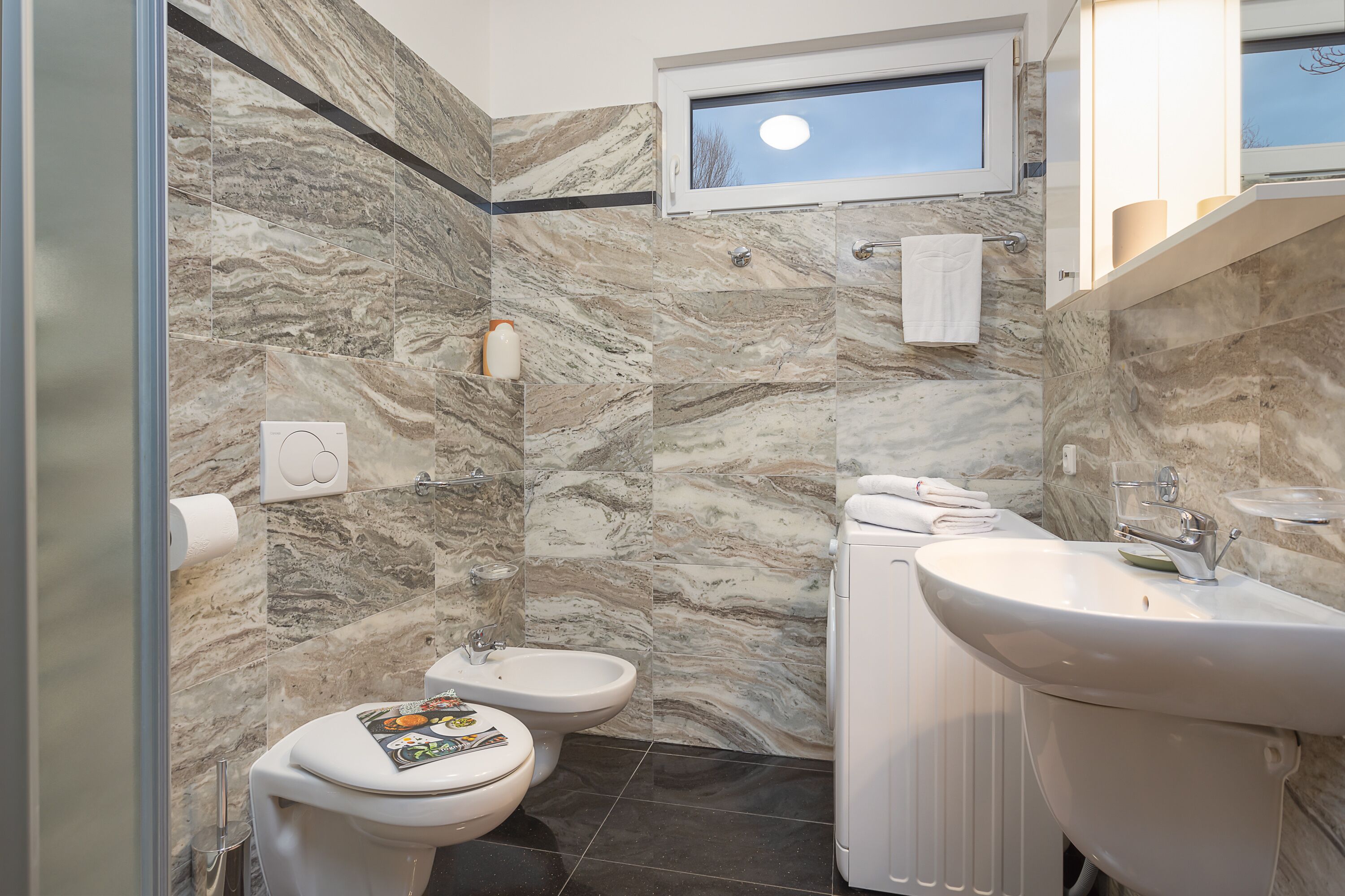 A bathroom with marble walls, a bidet, a toilet, a sink, and a window.