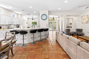 A modern kitchen with white cabinets, a central island with bar stools, a large clock, and a view into a dining area.