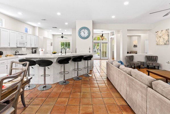 Cozinha moderna com armários brancos, uma ilha central com banquetas e um relógio grande, com vista para uma sala de jantar.