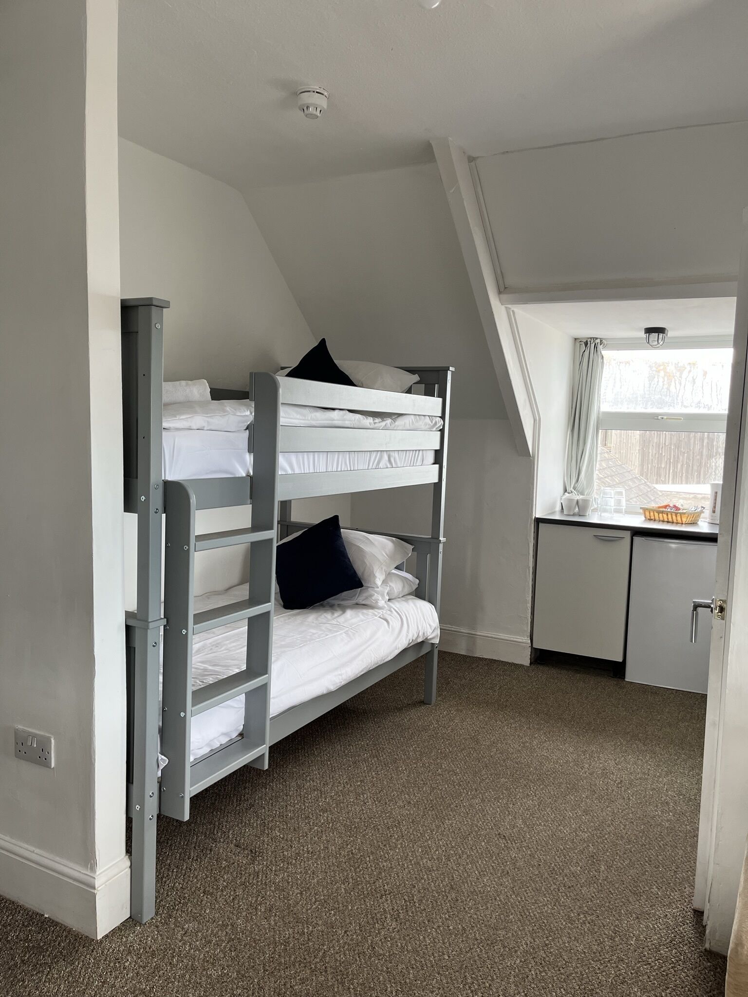 A dorm-style room with bunk beds, a kitchenette, and a window with curtains.