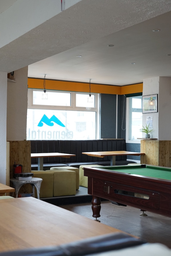 A billiards room with tables and a pool table, a window with a logo, and a seating area with a bench.