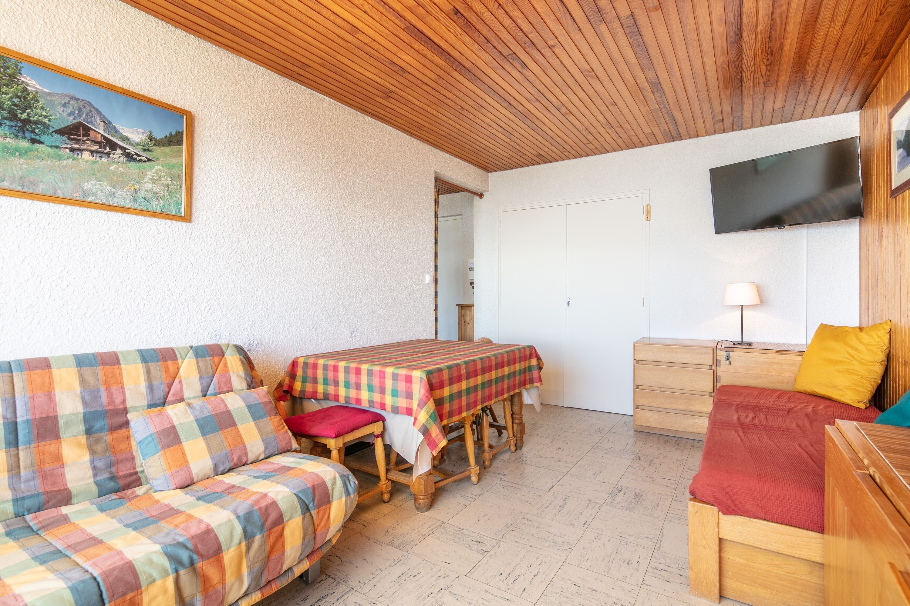 Een gezellige kamer met een houten plafond, een bank, een tafel met een geruit tafelkleed, een bed met een rode deken, een houten dressoir en een aan de muur bevestigde televisie.