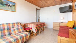 Ein gemütliches Zimmer mit Holzboden, einer Couch, einem Tisch mit kariertem Tuch, einem Bett mit roter Decke, einer Holzkommode und einem an der Wand befestigten Fernseher.