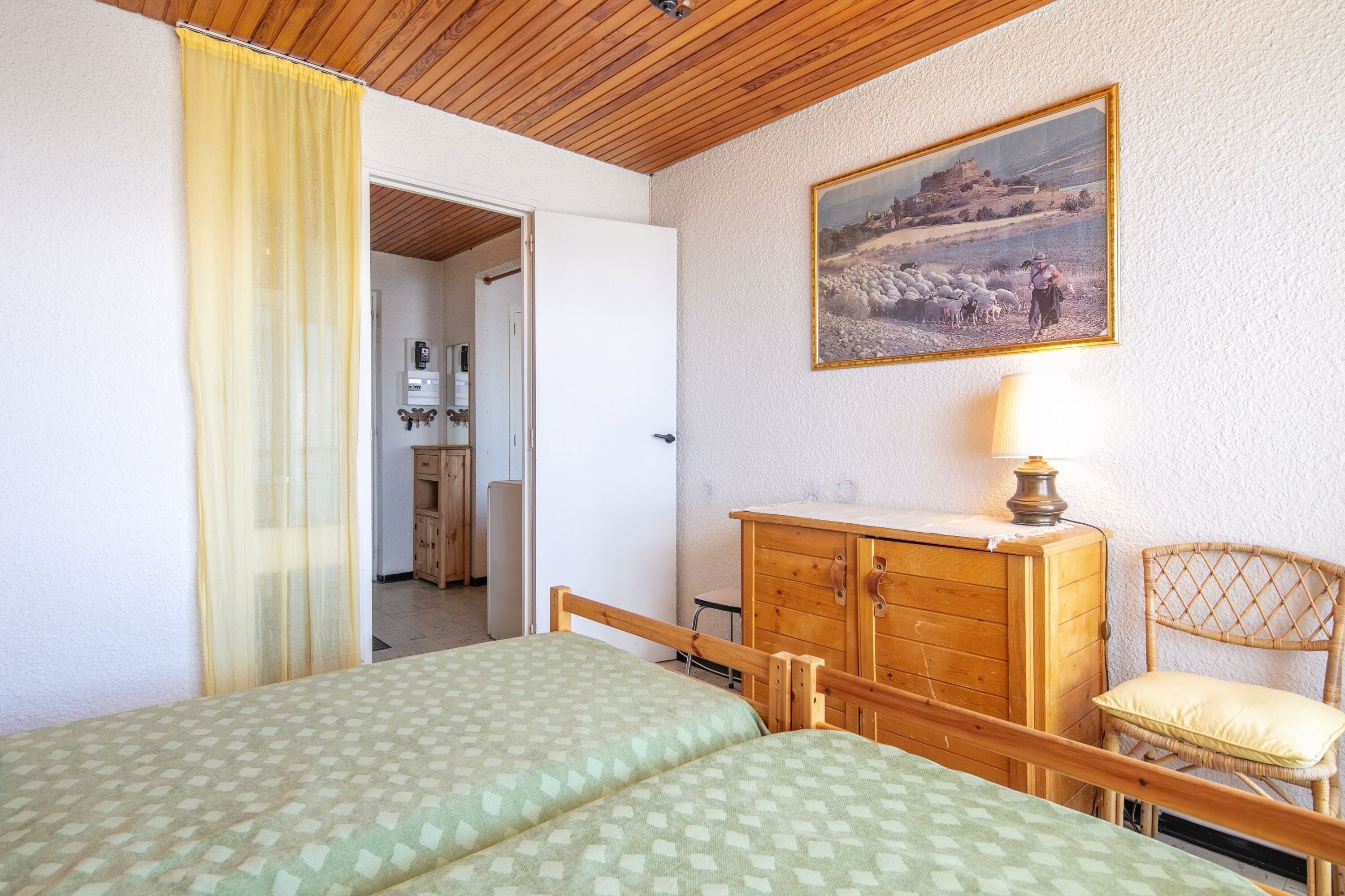 Een slaapkamer met twee bedden, een houten dressoir, een rieten stoel, een schilderij aan de muur en een deur naar een badkamer.