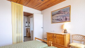 Ein Schlafzimmer mit zwei Betten, einer Holzkommode, einem Korbstuhl, einem Bild an der Wand und einer Tür zum Badezimmer.