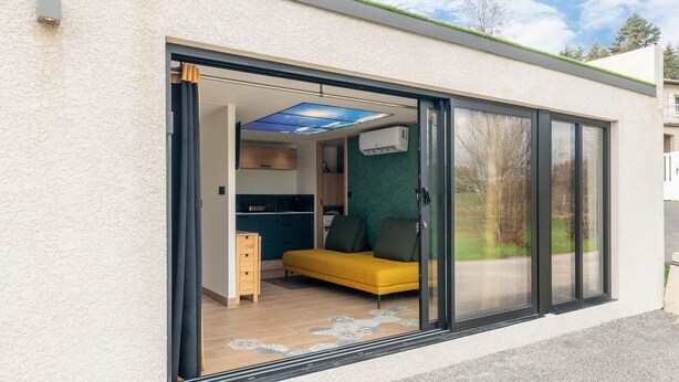 Una casa moderna con un cerramiento de vidrio corredera que da a una sala de estar con un sofá amarillo y una cocina a la vista.