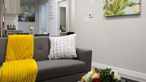 En stue med en grå sofa, et gult plaide og en hvid pude. Der er et maleri med blomster på væggen og en buket blomster på gulvet.