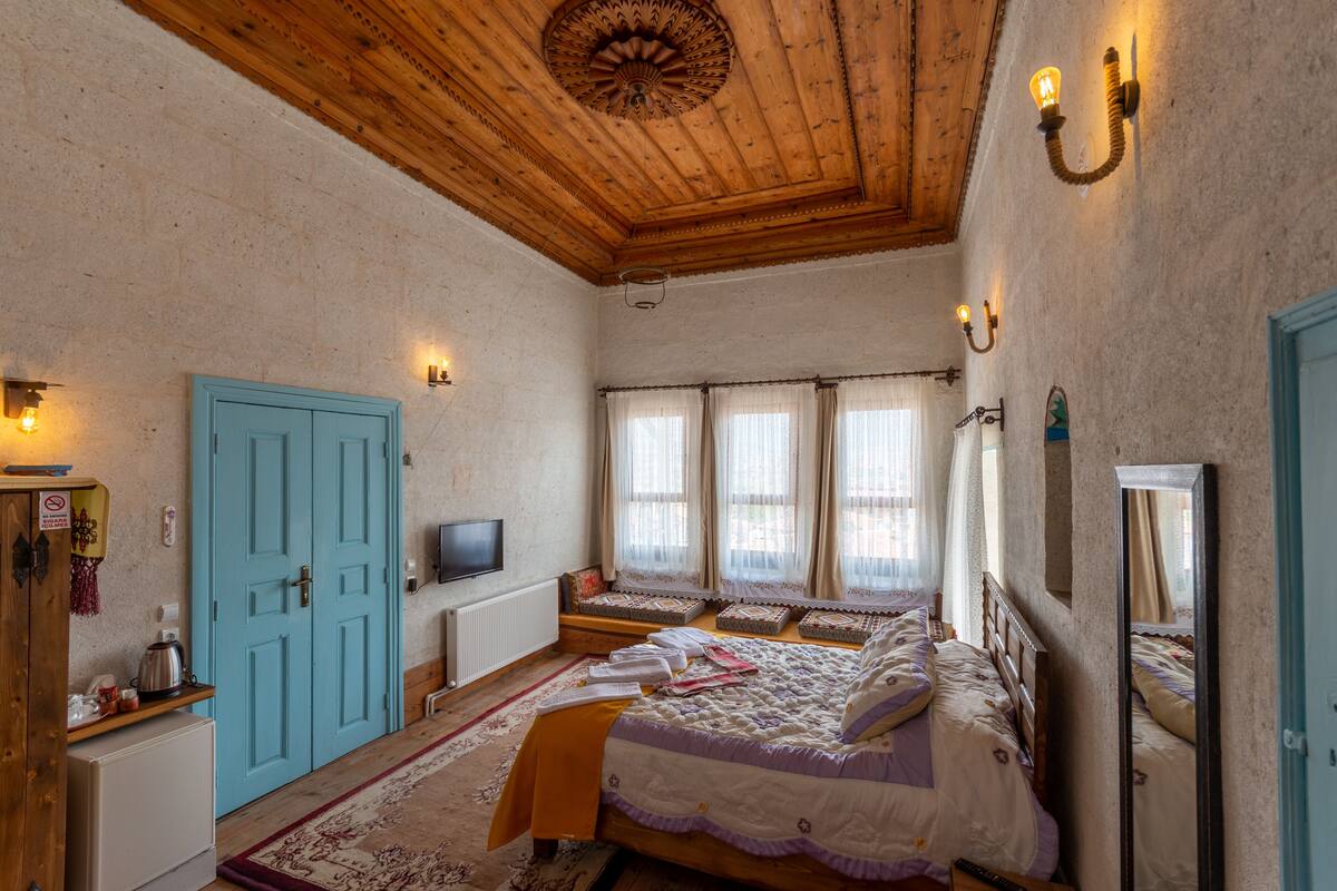 Een kamer met een houten plafond, een blauwe deur, een bed met een wit en paars dekbed, een kleine tafel met een waterkoker en een televisie aan de muur.
