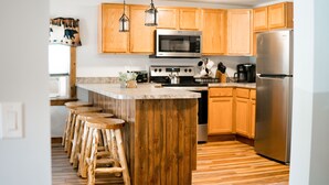 Una cocina con armarios de madera, un refrigerador de acero inoxidable, un microondas, una estufa y taburetes de madera.