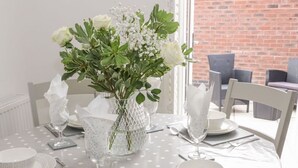 Une table à manger est dressée avec de l’argenterie, de la vaisselle blanche et des verres, et entourée de chaises blanches. Un vase rempli de fleurs blanches est posé sur la table.