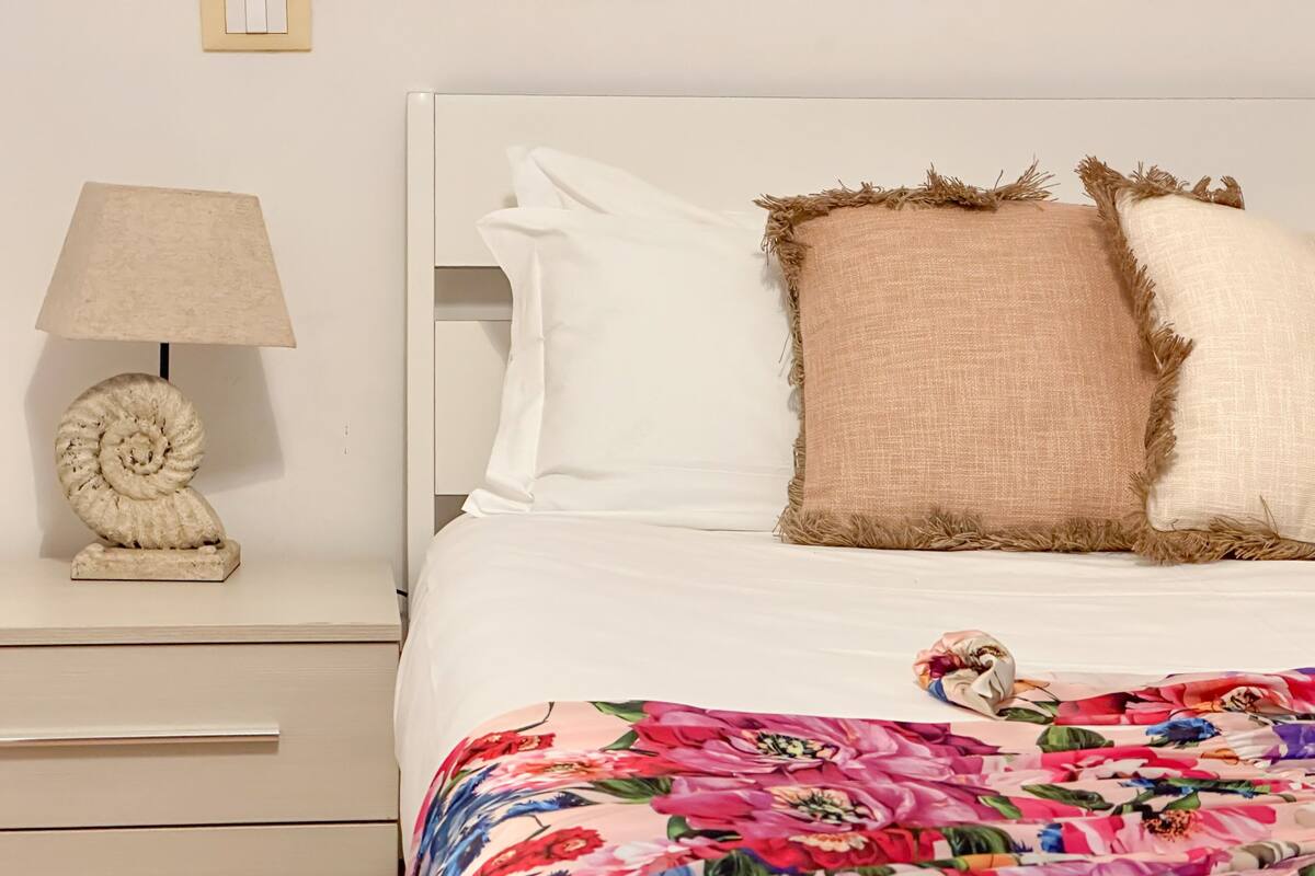 A neatly made bed with a floral blanket, two pillows, and a nightstand with a lamp and a decorative shell.