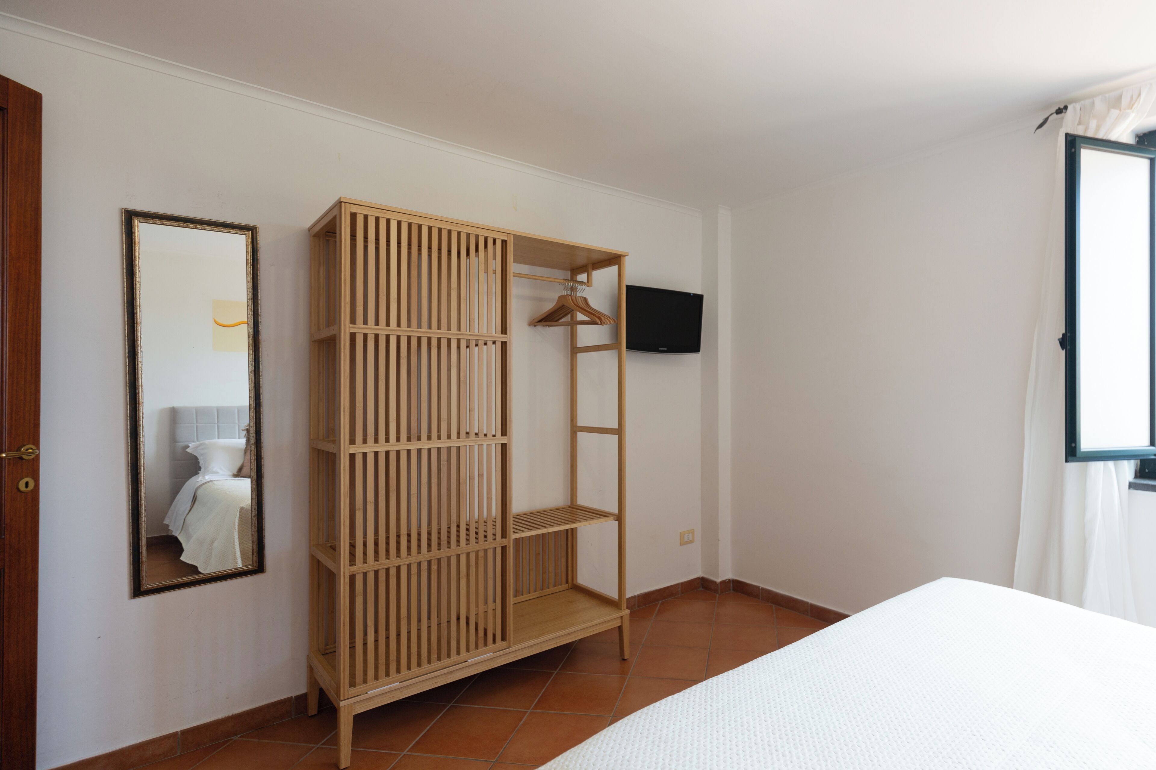 A bedroom with a bed, a wooden wardrobe, a mirror, and a TV on the wall.